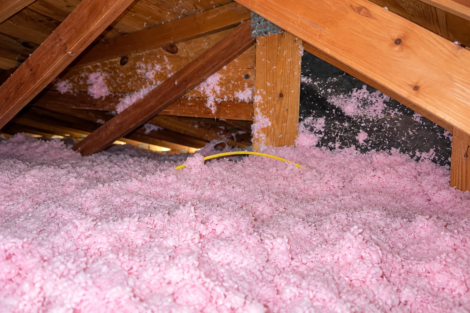 Pink loose-fill insulation covers an attic floor beneath exposed wooden roof trusses.
