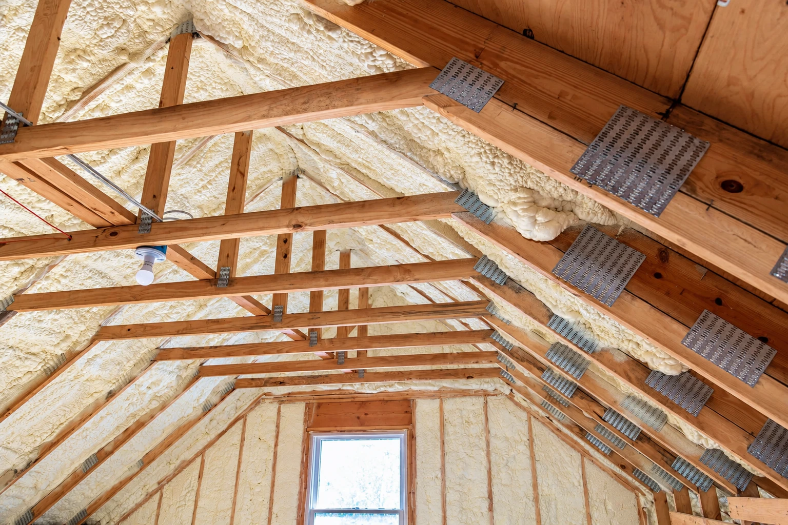 Closed-cell foam insulation coats the underside of a timber-framed roof, with metal connector plates across the rafters.