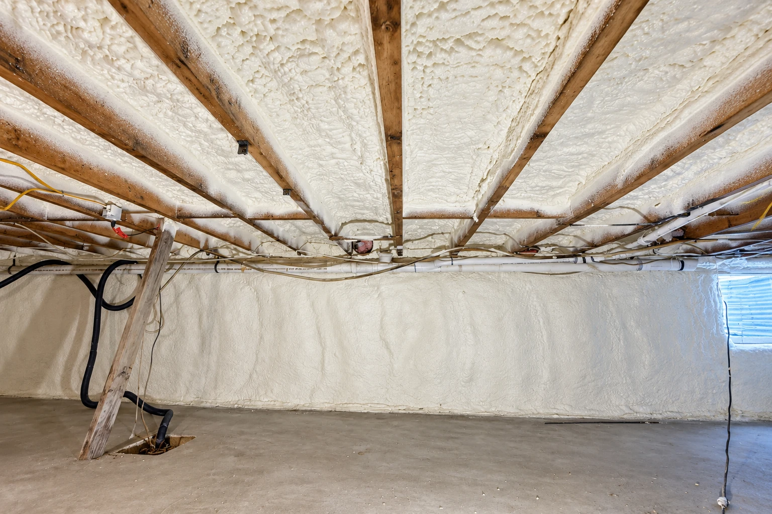 Crawl space ceiling and walls coated in white spray foam, with exposed wooden joists, pipes, and wiring.