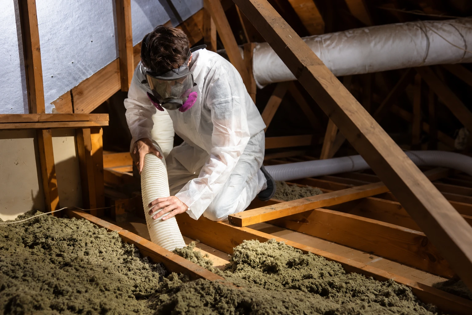 A worker wearing protective gear uses a vacuum hose to remove old insulation between attic joists.
