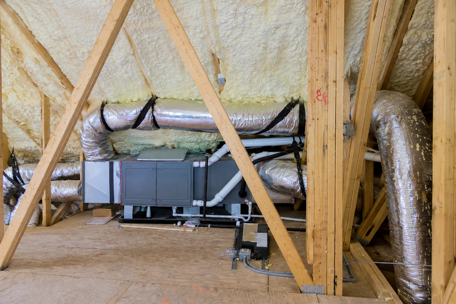 Open-cell foam insulation coats the roof framing in an attic with a central HVAC unit and silver ducts.