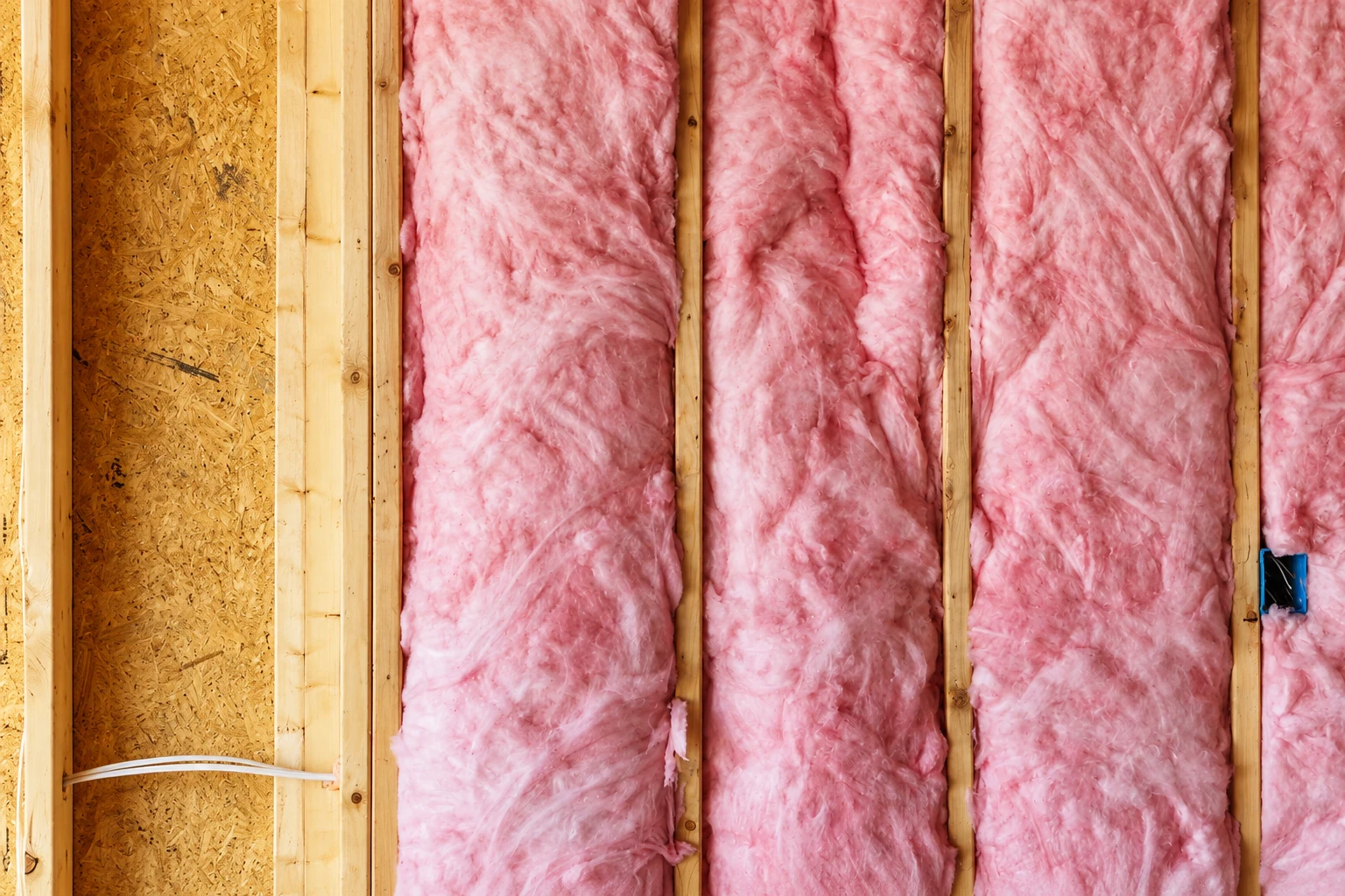 Pink fiberglass batts fill wooden wall cavities beside an exposed sheathed section and electrical outlet.