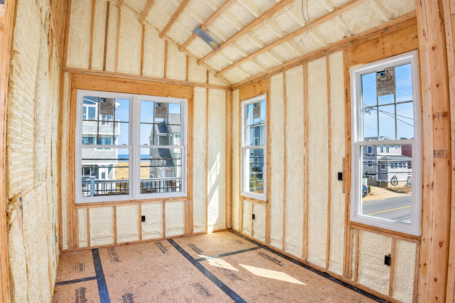 Spray foam insulation fills the wall and roof framing of an unfinished room with several windows.